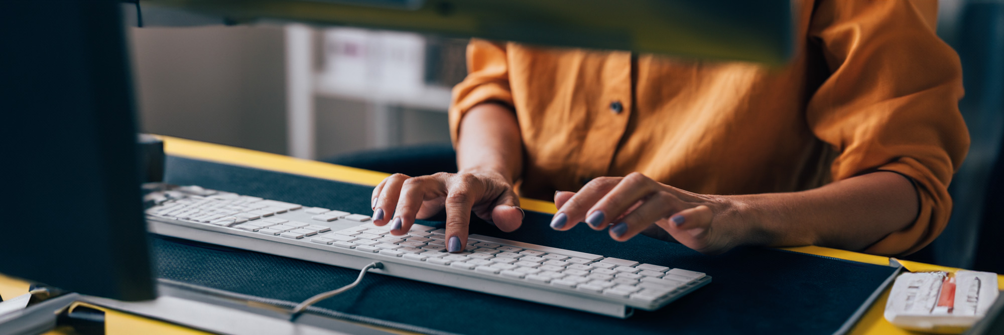 Close-up of a person with painted nails typing on a white keyboard at a desk with a yellow surface.