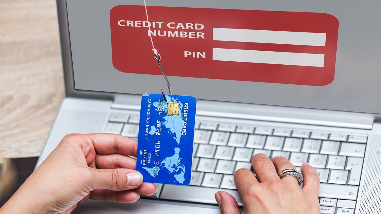 A person holding a credit card caught on a phishing hook in front of a fake login screen on a laptop, symbolizing online fraud or identity theft.