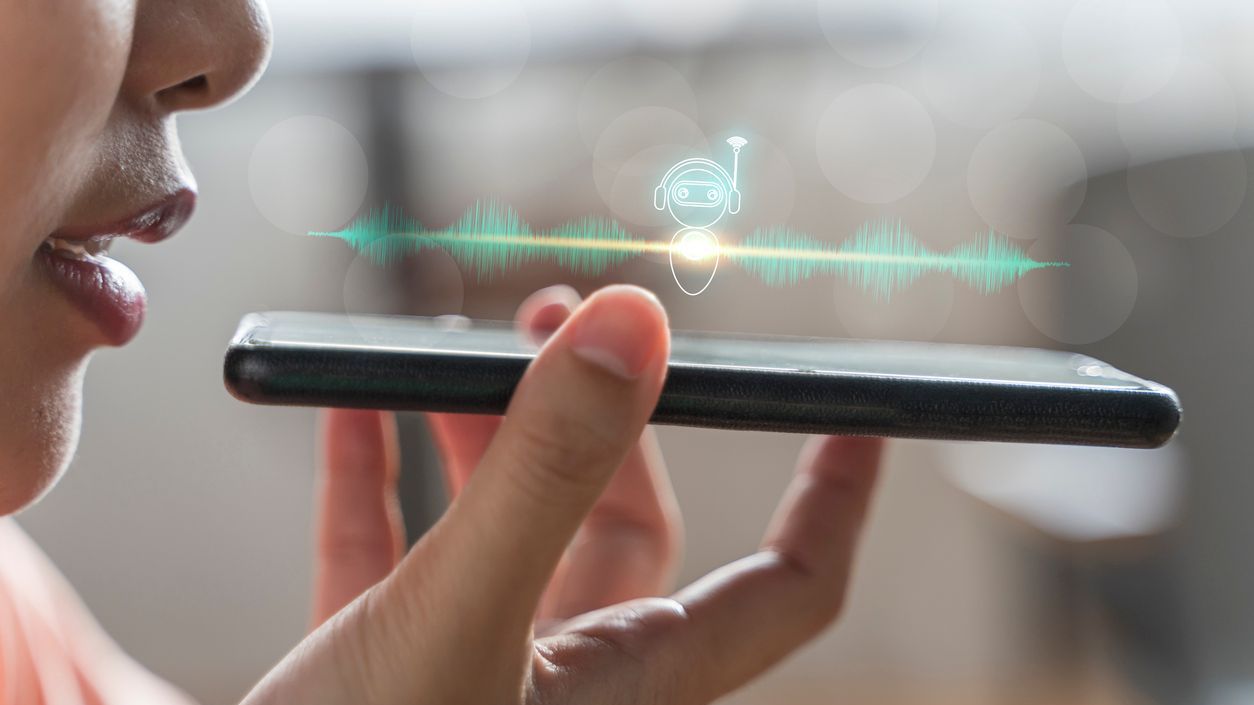 A person speaking into a smartphone with a digital voice assistant icon and soundwave graphic displayed, representing voice recognition technology.