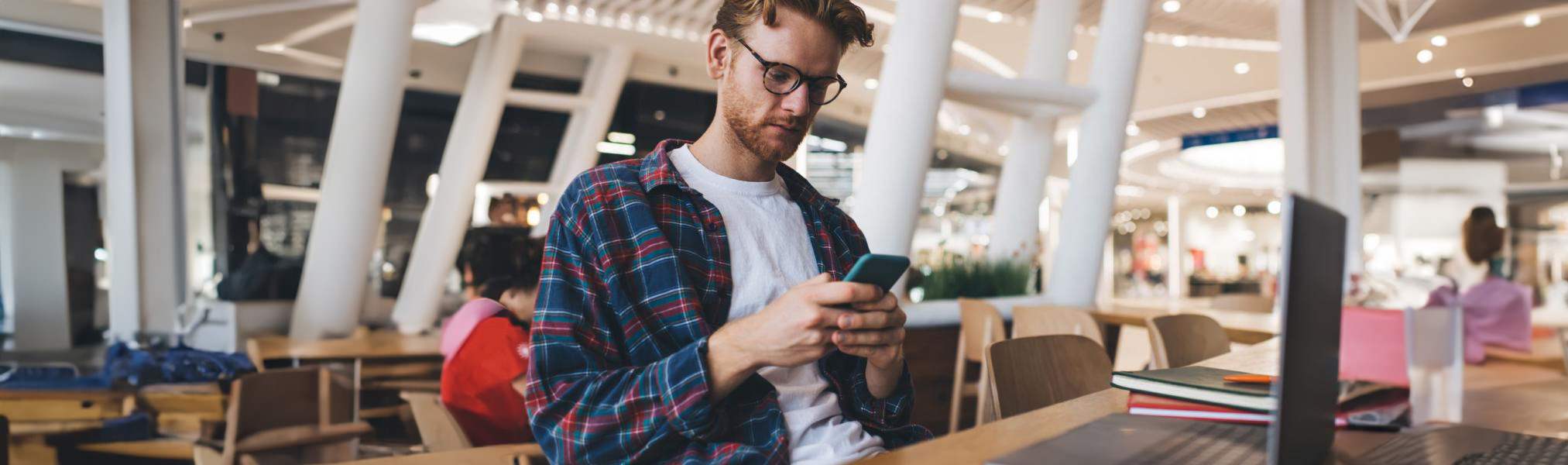 Jeune homme portant des lunettes et une chemise à carreaux utilisant un smartphone à une table avec un ordinateur portable et des cahiers dans un cadre intérieur moderne.