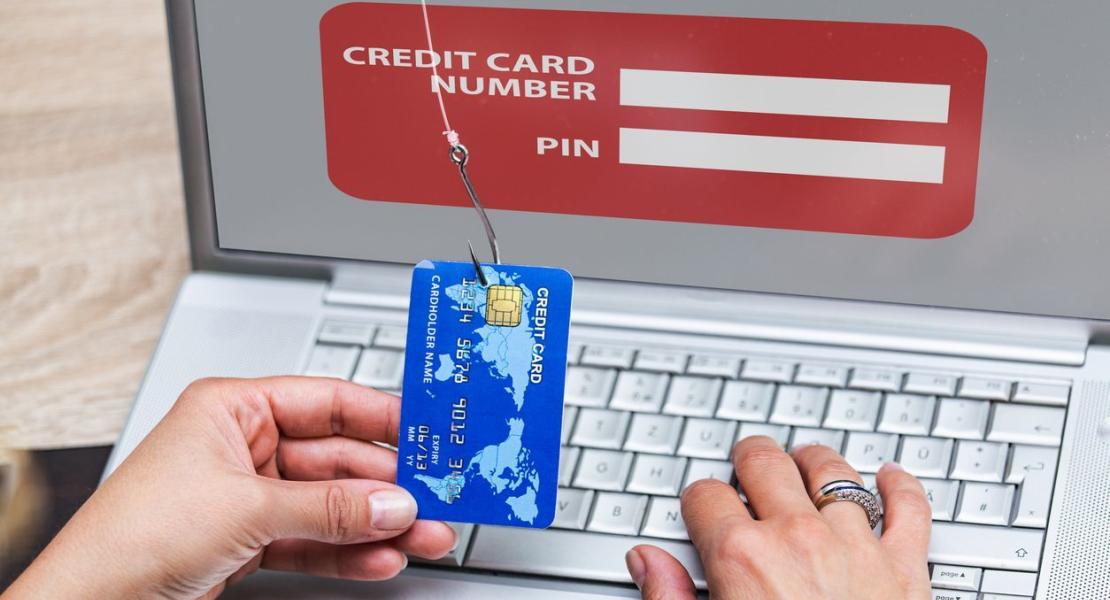 A person holding a credit card caught on a phishing hook in front of a fake login screen on a laptop, symbolizing online fraud or identity theft.