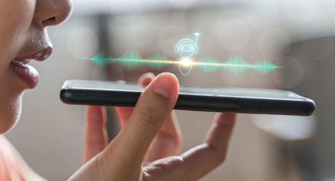 A person speaking into a smartphone with a digital voice assistant icon and soundwave graphic displayed, representing voice recognition technology.