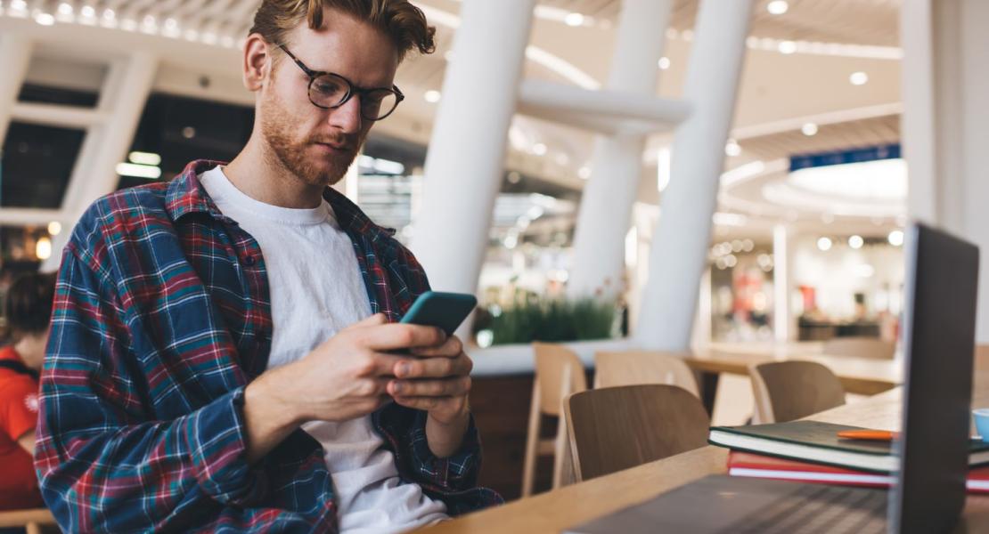 Young man wearing glasses and a plaid shirt using a smartphone at a table with a laptop and notebooks in a modern indoor setting.