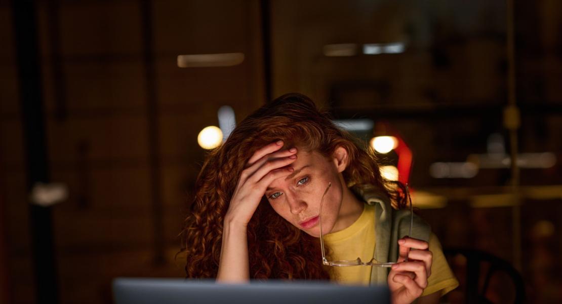 Femme frustrée avec des cheveux roux et bouclés, tenant des lunettes et regardant l'écran d'un ordinateur portable dans un bureau faiblement éclairé.