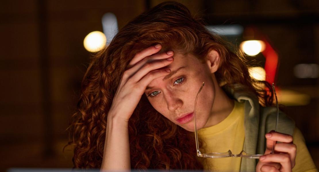 Frustrated woman with red curly hair holding glasses and looking at a laptop screen in a dimly lit office.